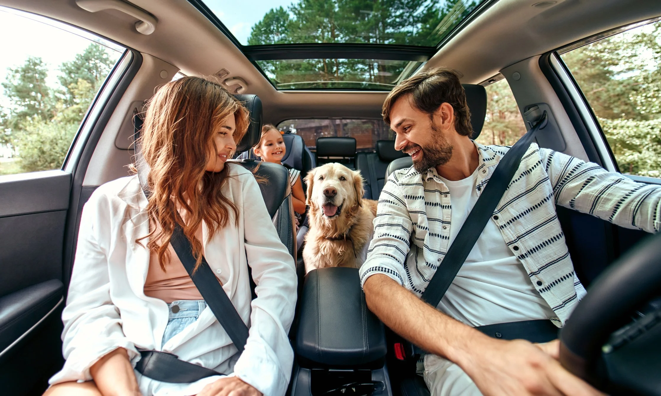 A family driving together, two adults in front, a child and a golden retriever in the back seat, everyone belted in.