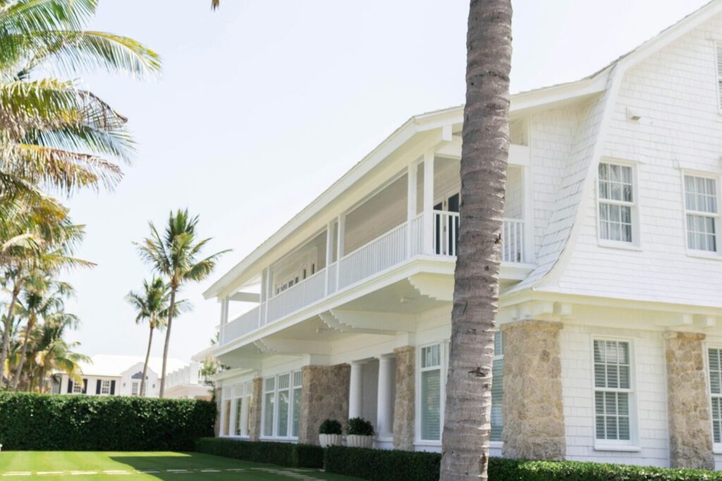A white two-storey Florida house behind palm trees and a clipped hedge